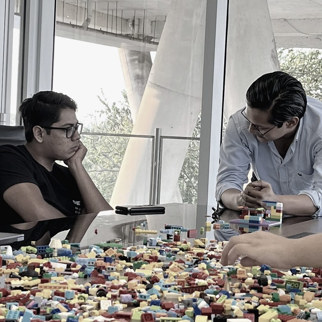 Dos integrantes de un equipo frente a una mesa cubierta de piezas: uno observa, el otro construye un modelo.